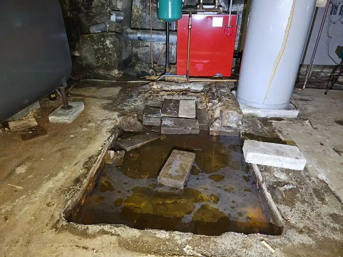 Water-filled hole in basement floor with bricks. Boiler, tank, and oil tank in the background.
