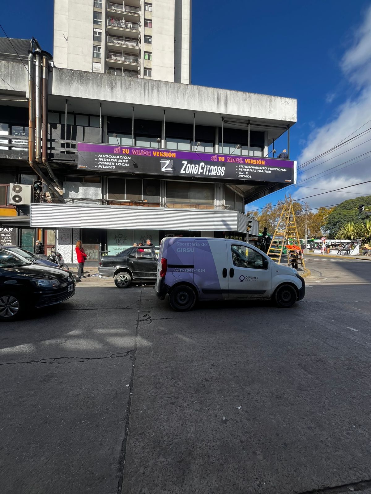 Escena callejera con un edificio y vehículos estacionados. Una furgoneta con un logotipo está en la calle.