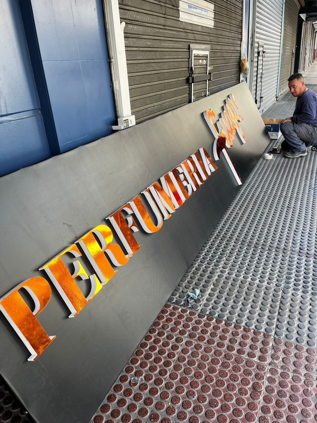 Un hombre instala un letrero que dice “Pier Fume” con letras de color naranja metálico. El letrero está en una pasarela.