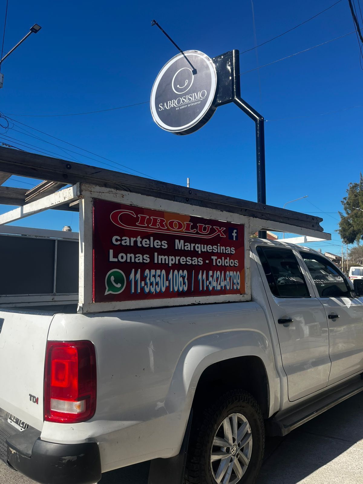 Camioneta blanca con un letrero publicitario en el techo y otro letrero. Cielo azul.