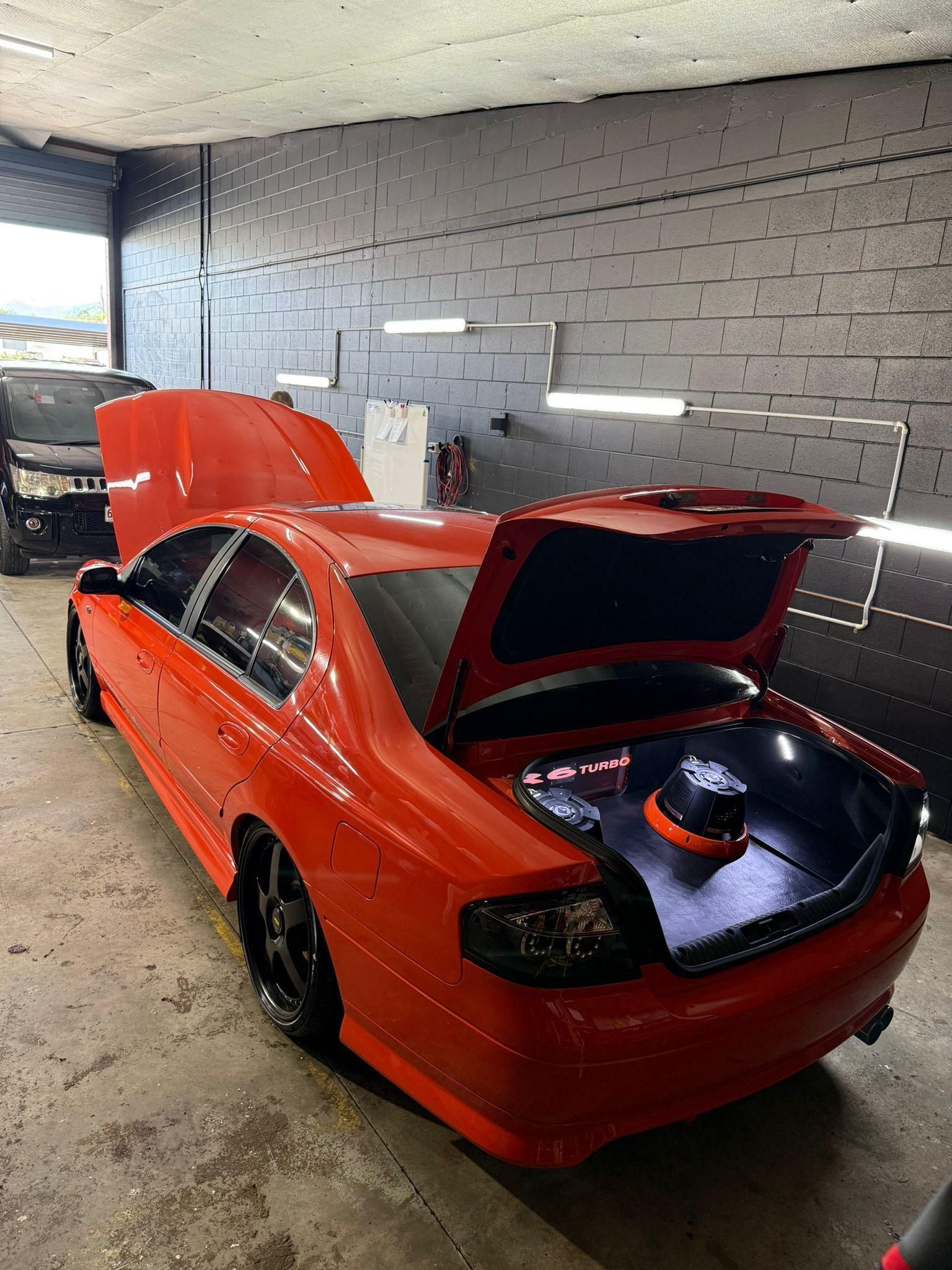 Red sports car in a garage with hood and rear hatch open, showing an audio setup in the trunk — Mobile Club Cars Cairns in Earlville, QLD