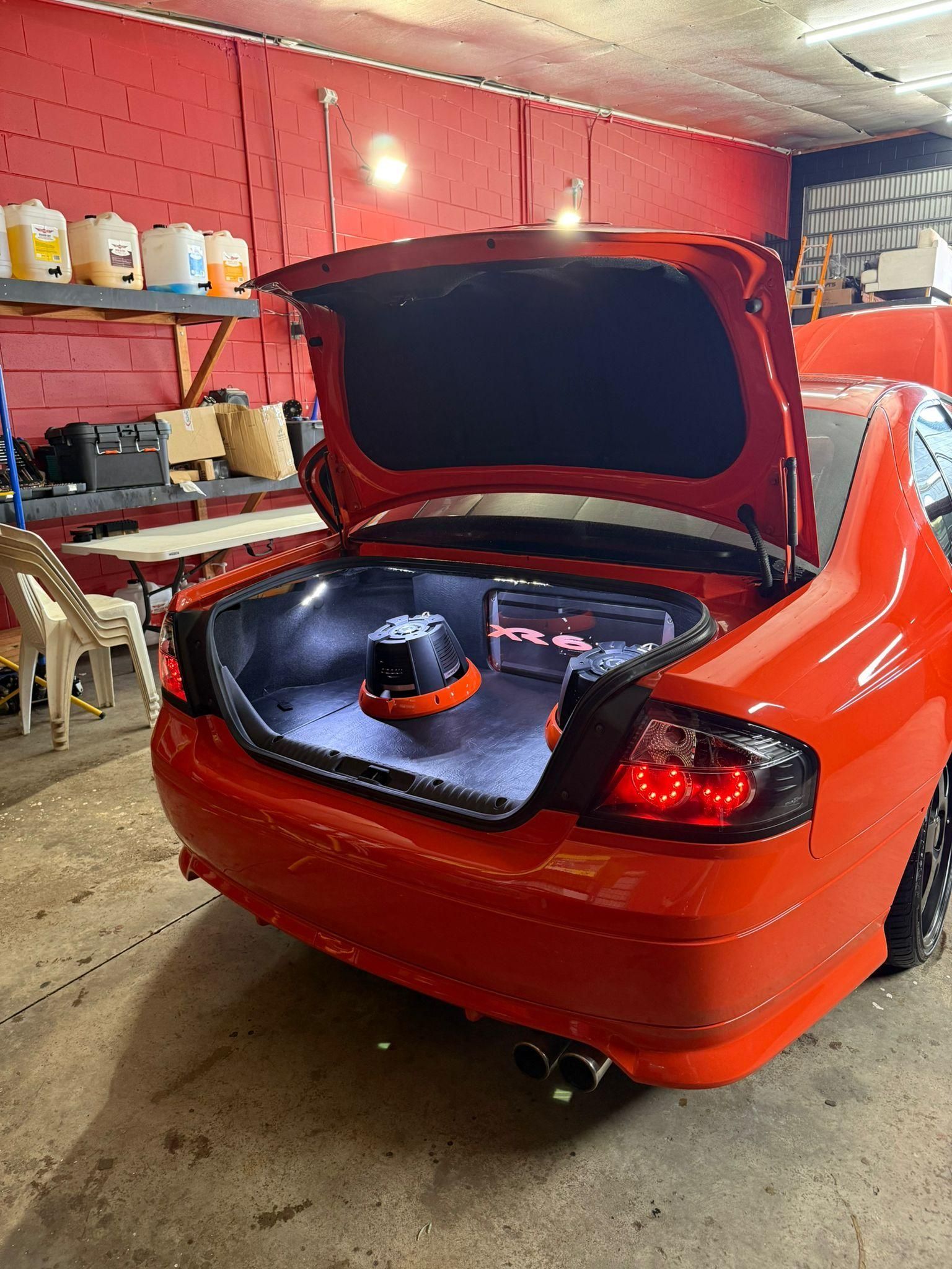 Orange sports car with trunk open in a garage, showing a large subwoofer setup in the back — Mobile Club Cars Cairns in Earlville, QLD