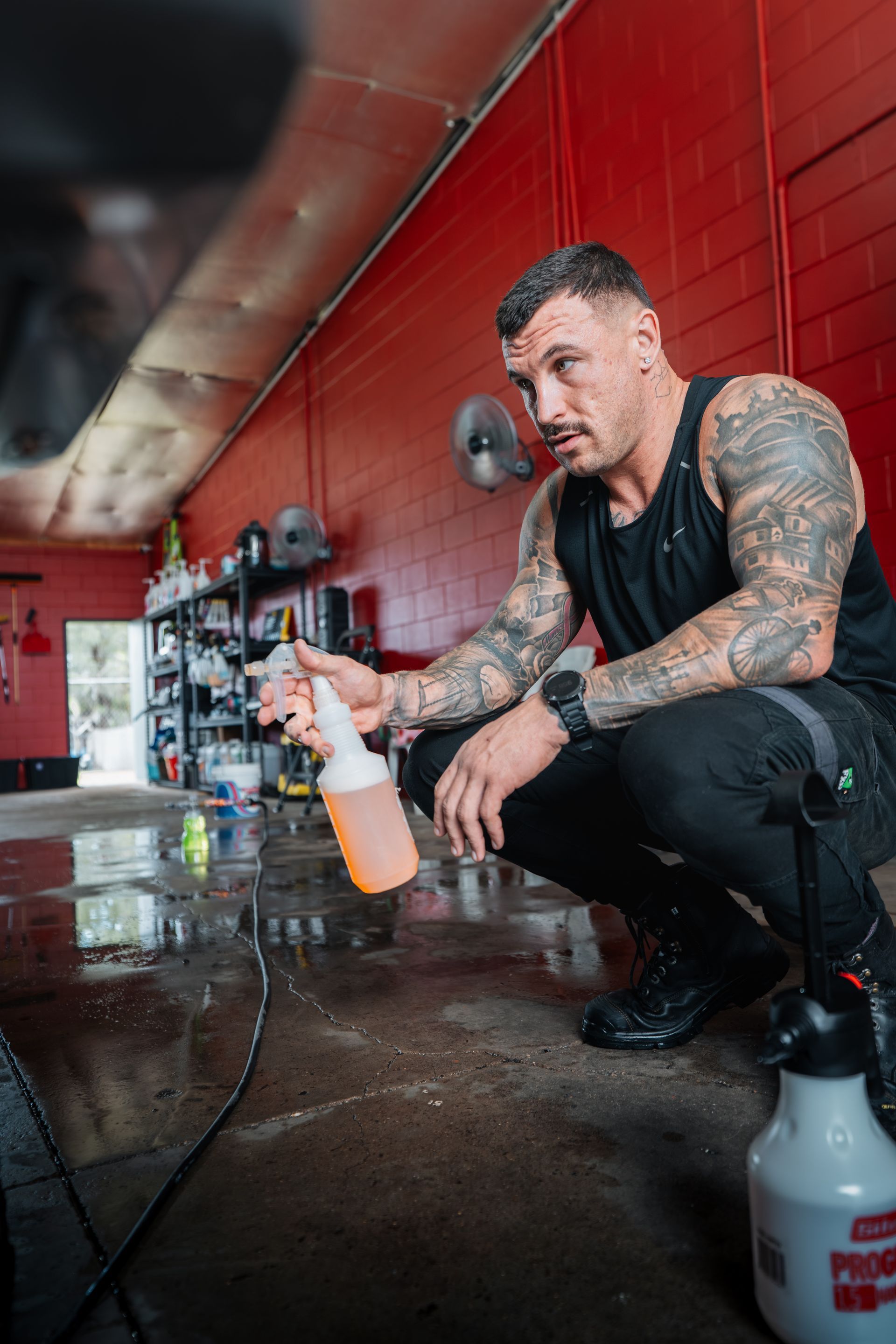 Man With Tattoos Crouching, Holding Spray Bottle in Auto Shop — Mobile Club Car Cleaning in Earlville, QLD