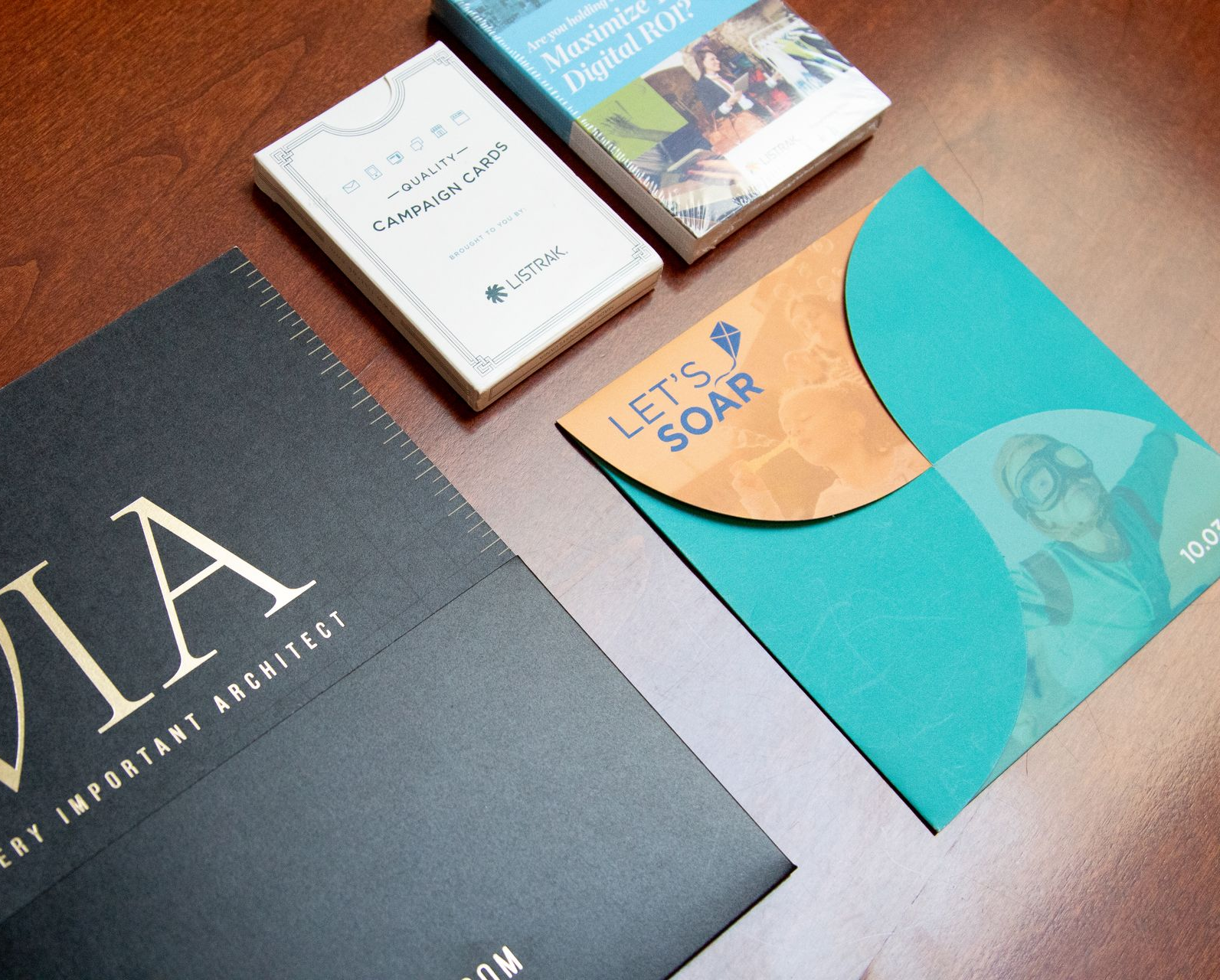 Books and envelopes on a brown surface. Items include a black folder with gold text and a teal and orange envelope with