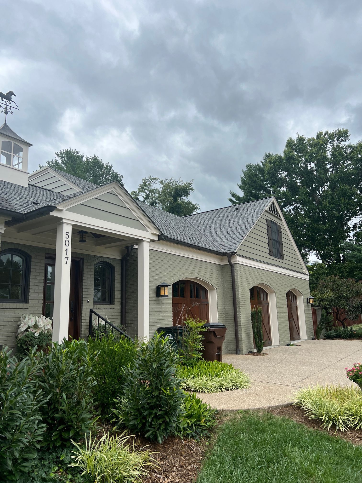 A light green house with a gray roof, dark garage doors, and a cobbled driveway under a cloudy sky.