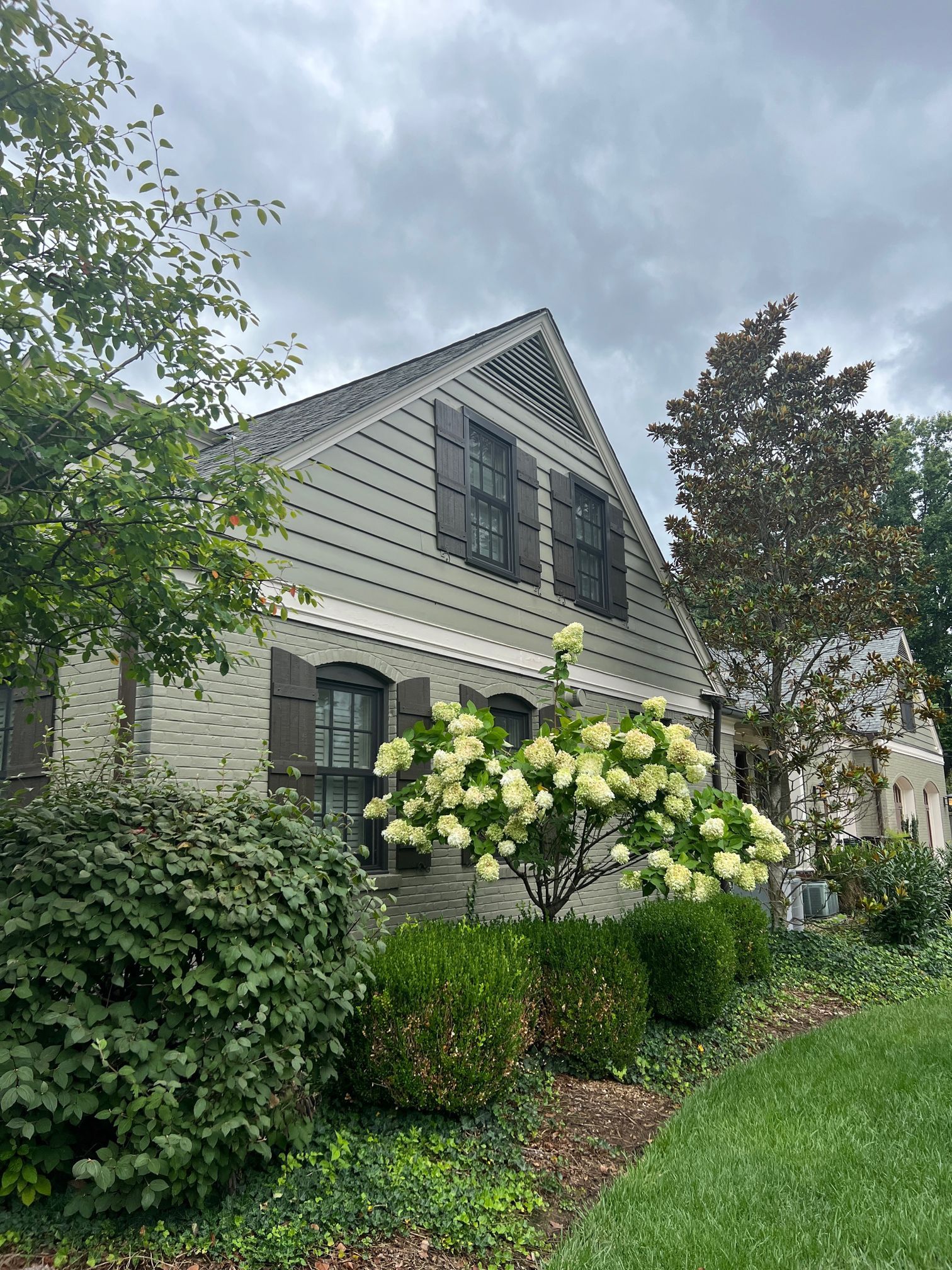 Gray house with dark shutters, lush landscaping under cloudy sky.