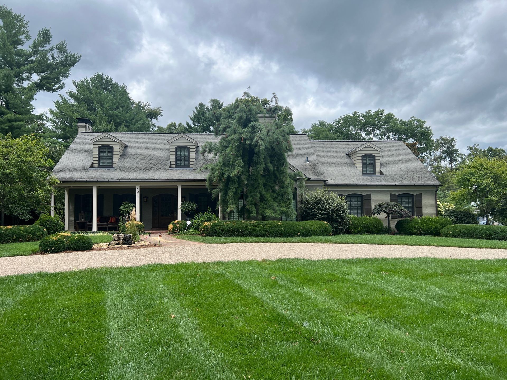 A house with a long porch and gravel driveway, surrounded by green grass and trees, under a cloudy sky.