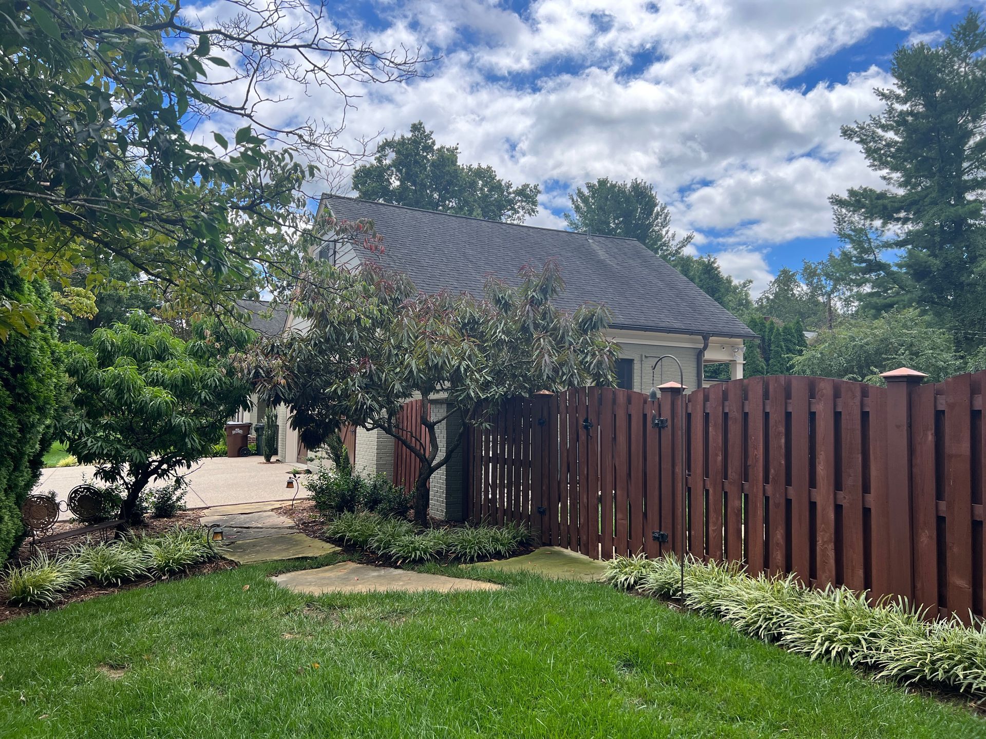 Lawn with brown fence, small tree, and house under a partly cloudy sky.