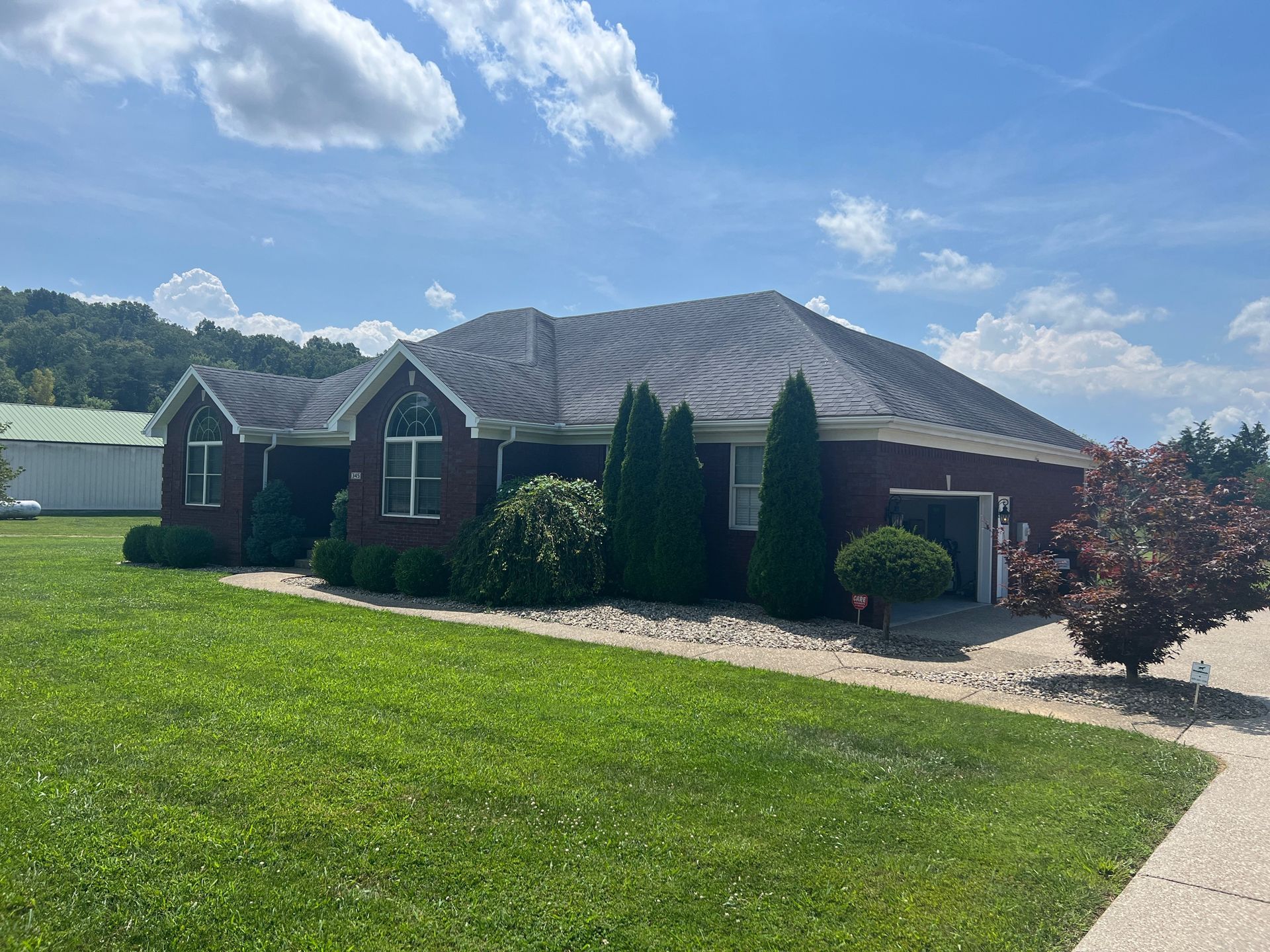 Brick house with dark roof, surrounded by green grass and trees, under a partly cloudy sky.