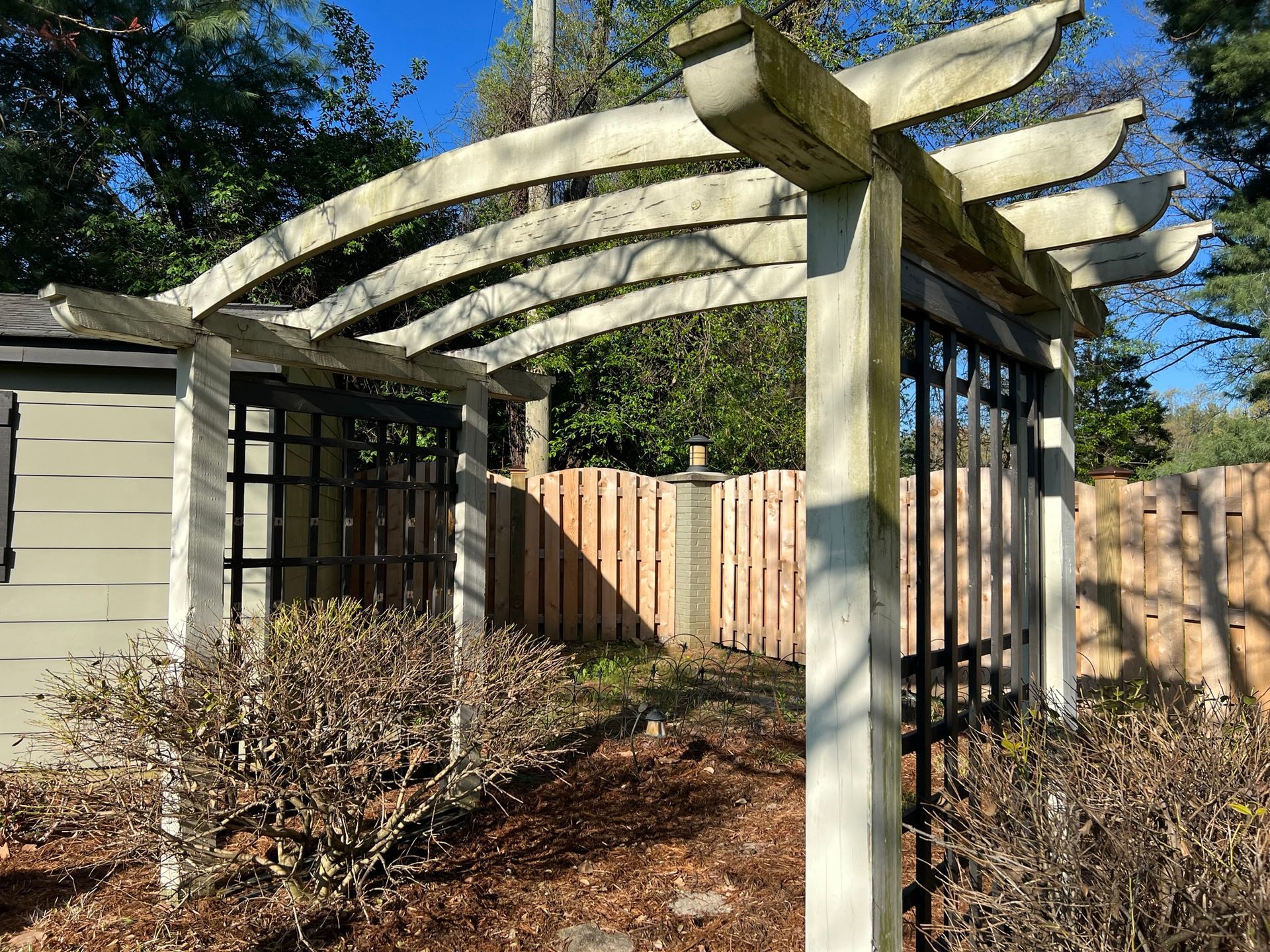 Wooden pergola over garden entrance with brown fence, shed, and shrubbery.