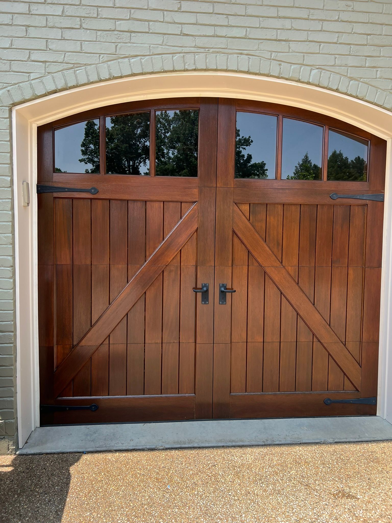Wooden garage door with cross braces and arched windows, against a brick building.