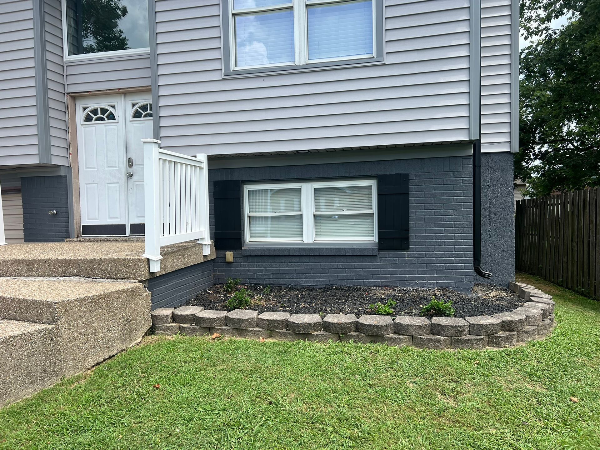 Two-story house with gray siding, a white door, and a black-painted brick foundation with a small flower bed.