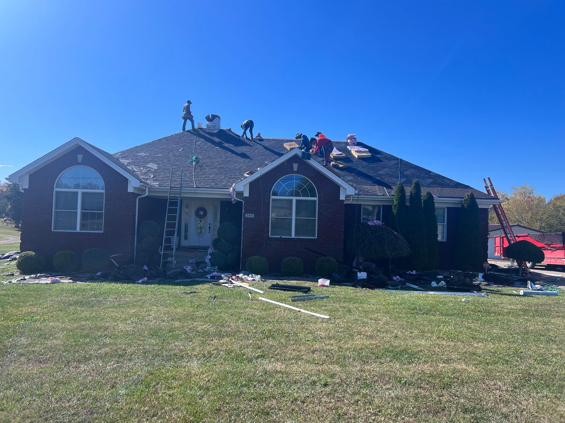 Roofers working on a brick house with blue sky. Debris and equipment are on the ground.