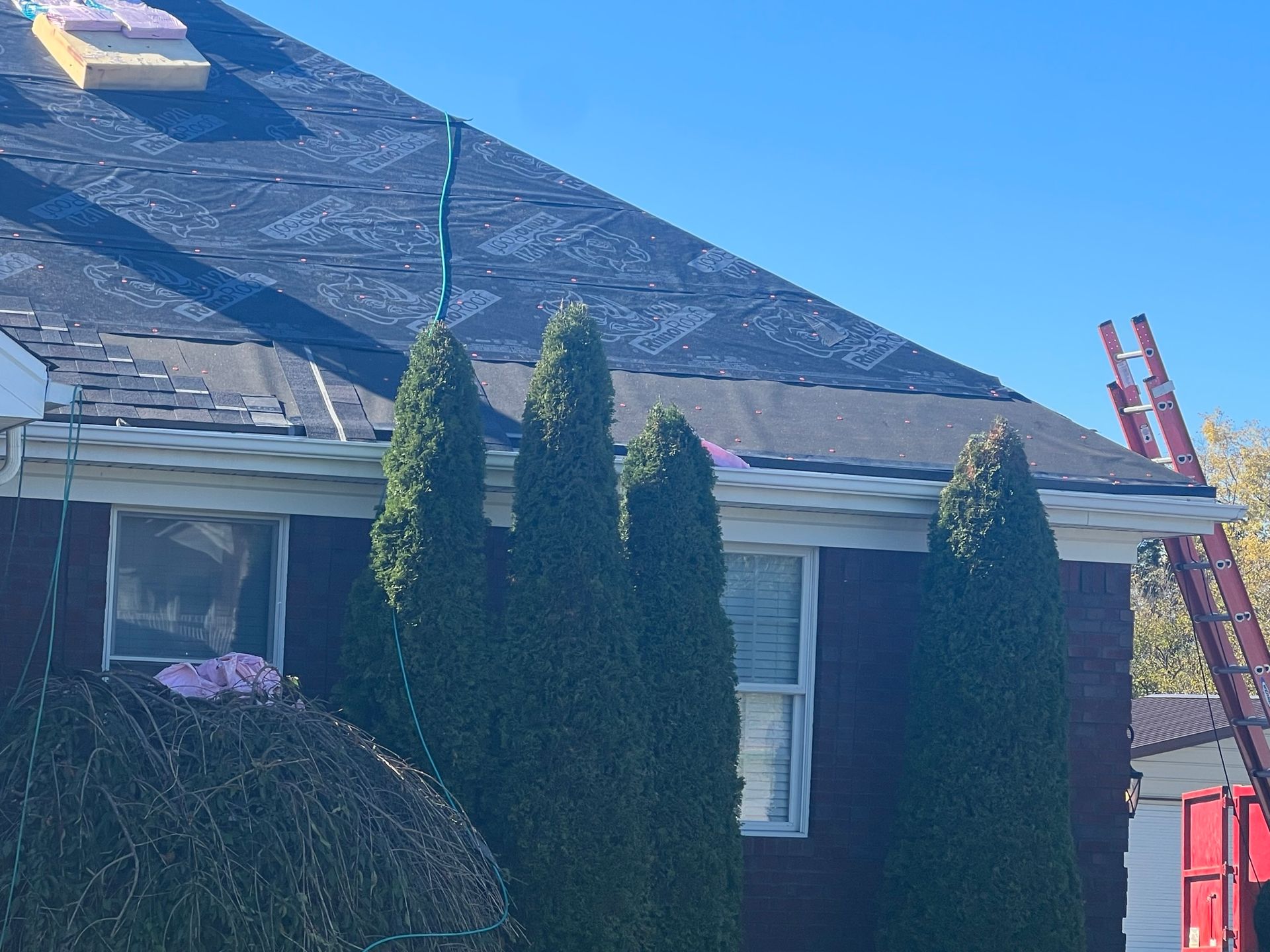 A house with a partially replaced roof. Green trees are in front, ladders on the side, and clear blue sky.