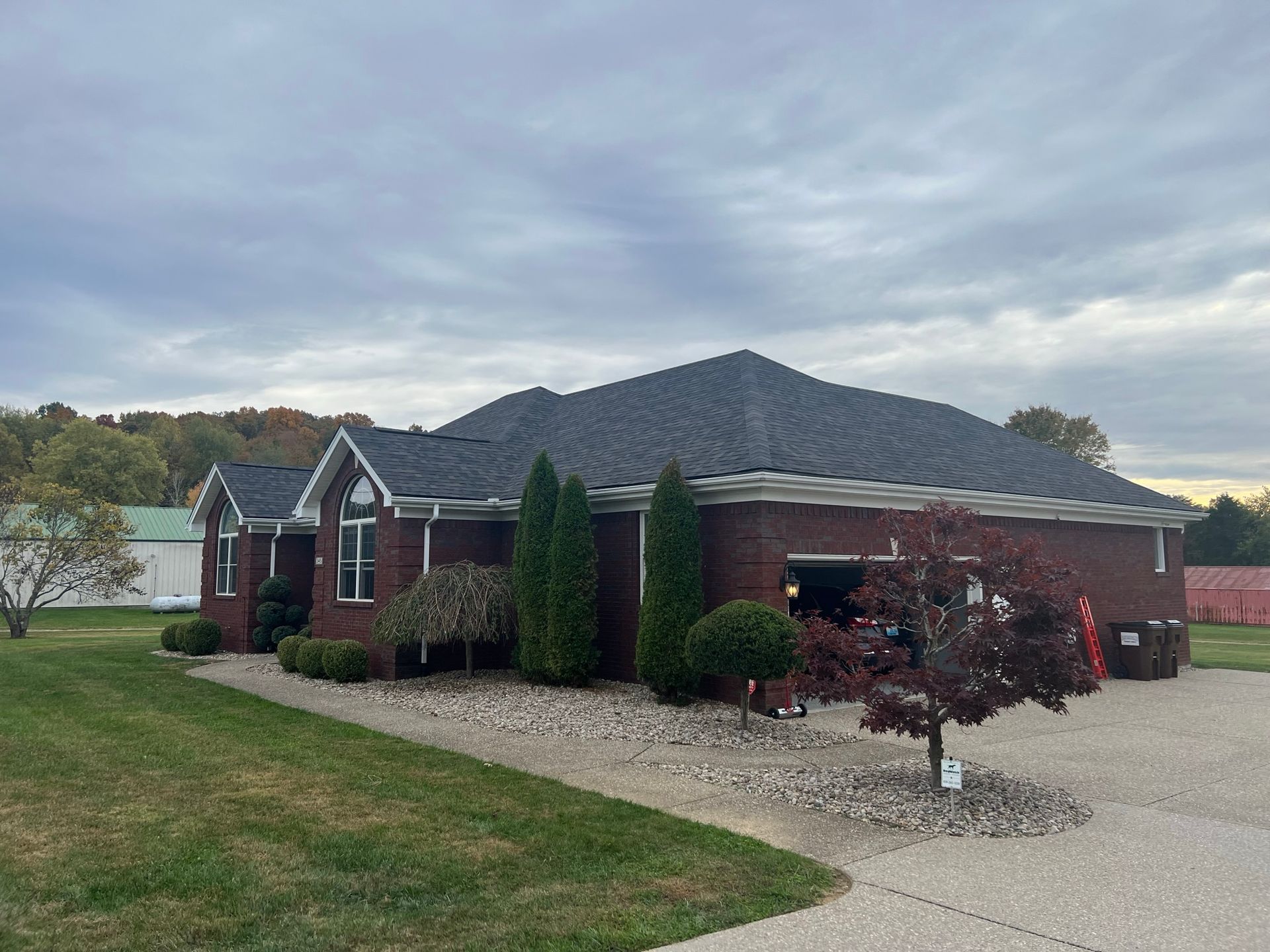 Red brick house with dark gray roof, evergreen shrubs, and red tree in front of a gravel driveway.