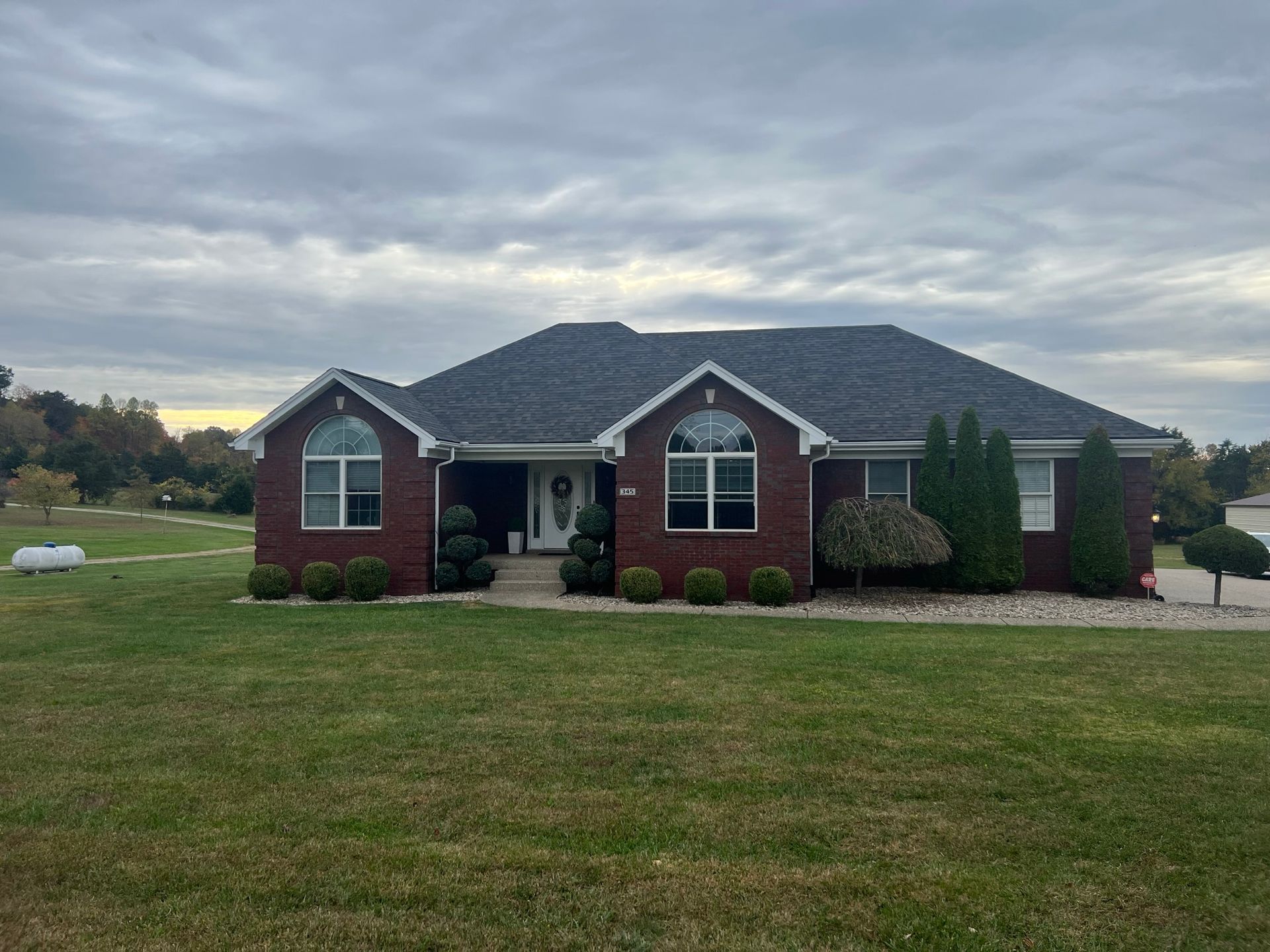 Red brick ranch-style house with dark roof, arched windows, and manicured landscaping on a cloudy day.