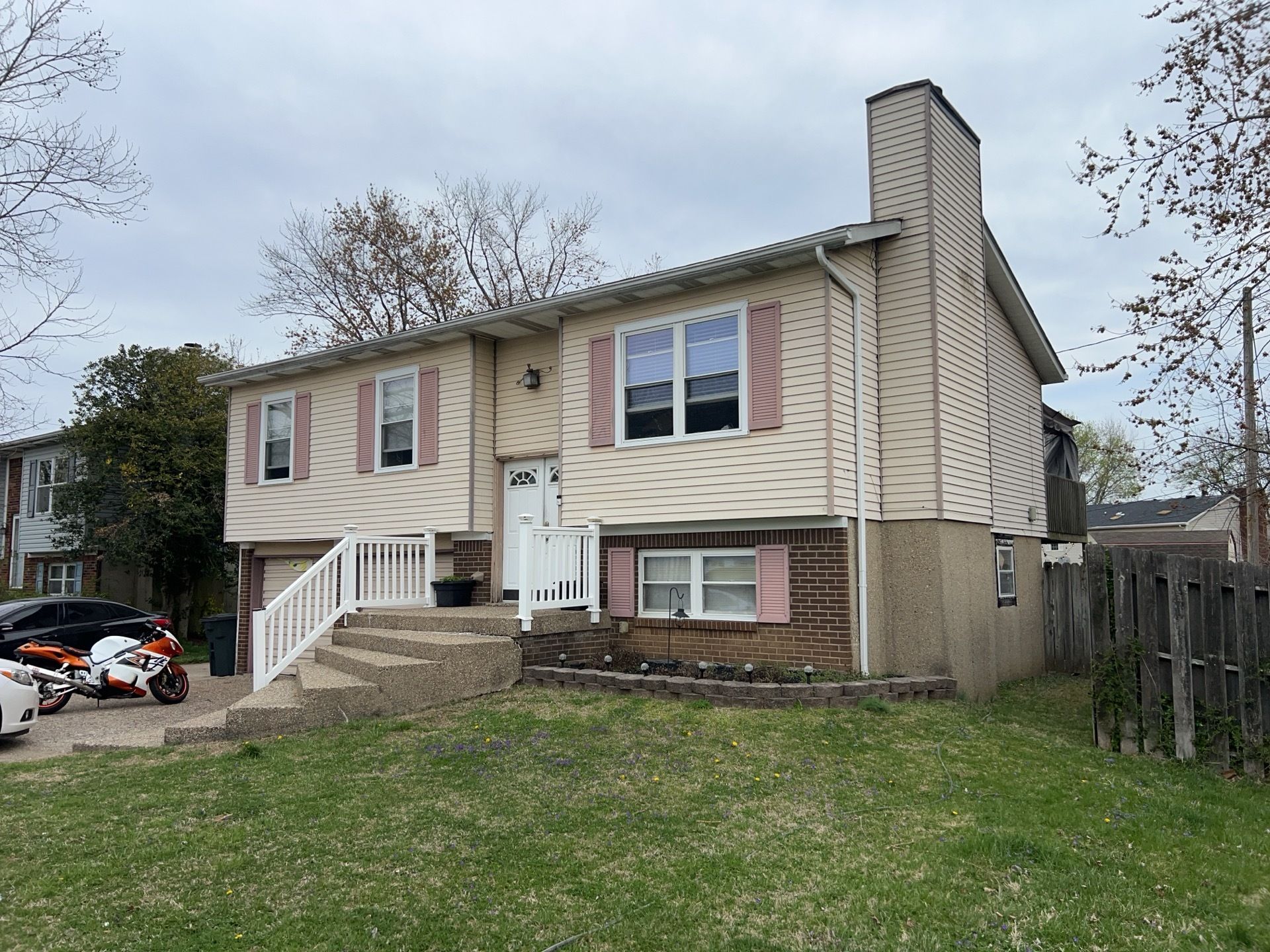 Tan split-level home with pink shutters and a concrete porch. A car and motorcycle are parked in the driveway.
