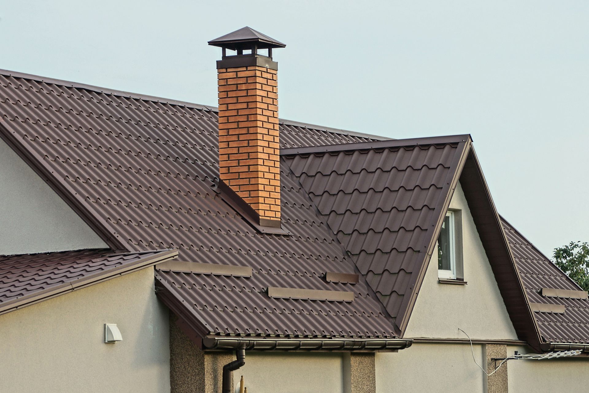 One large red brick chimney on a brown tiled roof of a brown house with a window against the sky