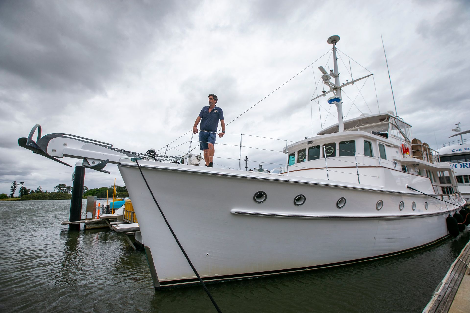 Man stands on the bow of a white yacht at a dock. Overcast sky.