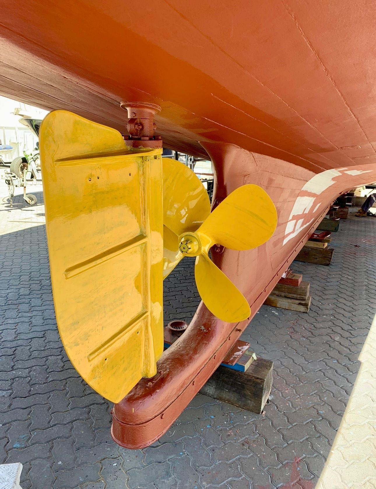 Yellow propeller and rudder on a red-painted ship hull, resting on blocks.