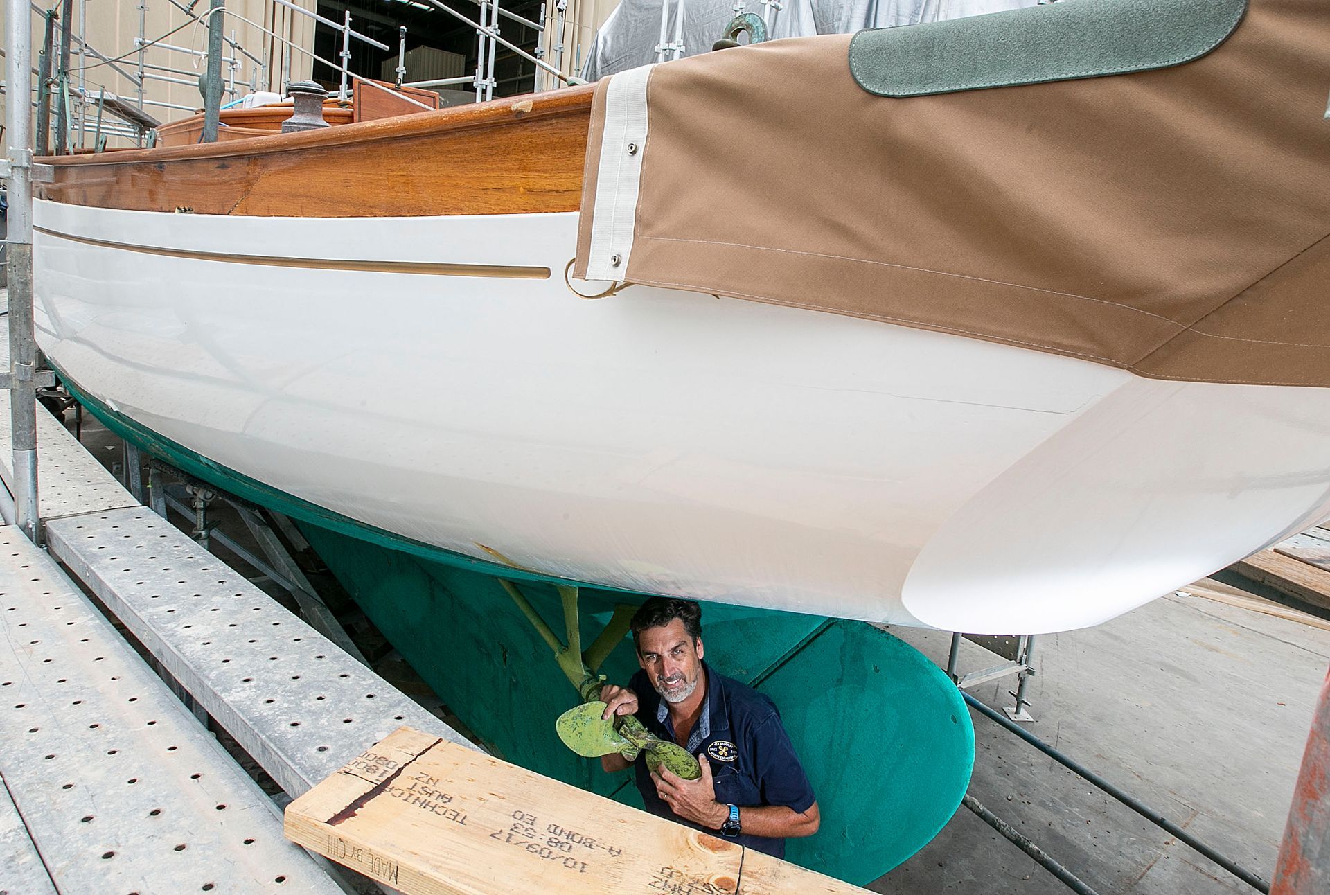 Man under a sailboat hull, holding a propeller. The boat is white with a green keel.