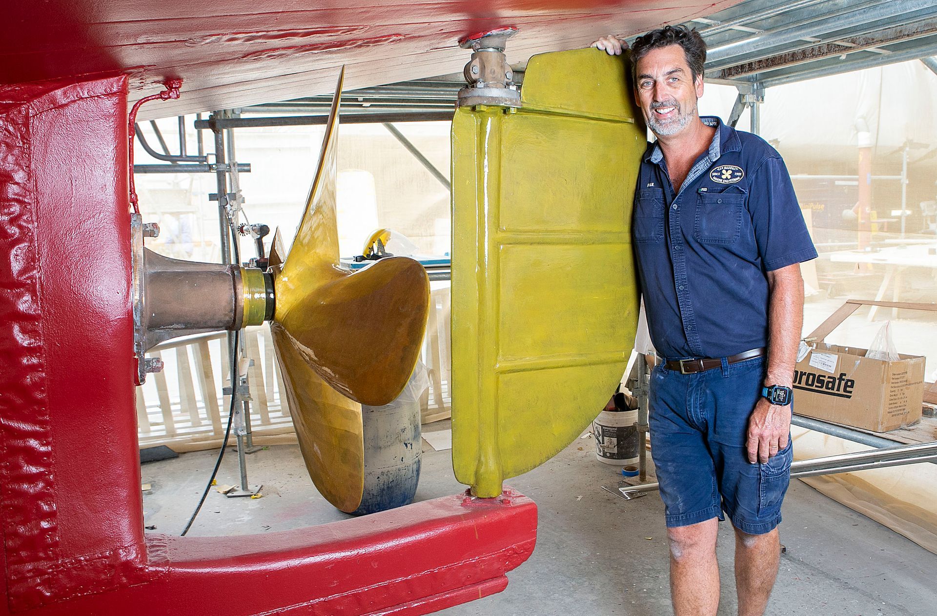 Man stands next to large, gold ship propeller and yellow rudder; red hull in a shipyard.