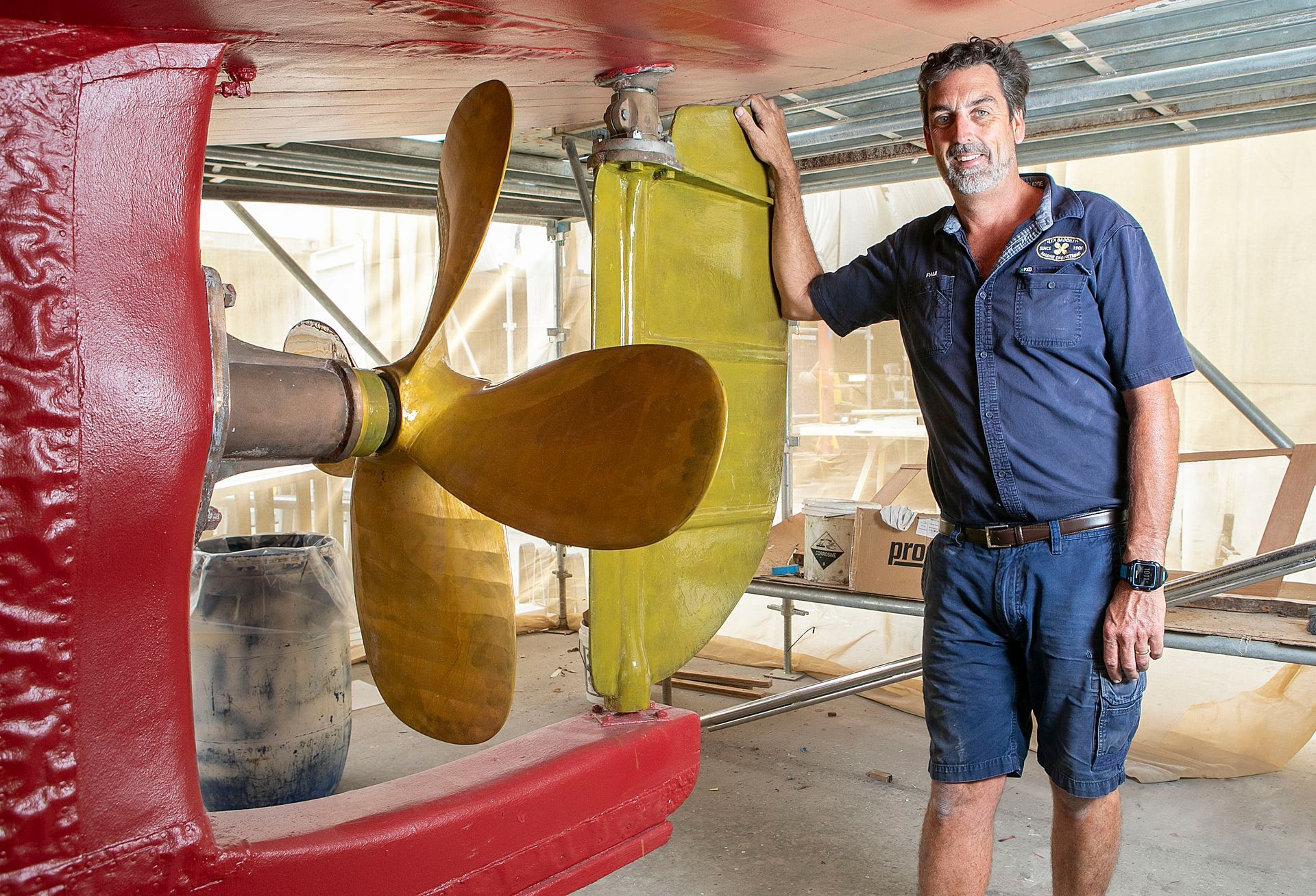 Man stands next to a boat propeller in a boat repair shop, red hull, yellow propeller.