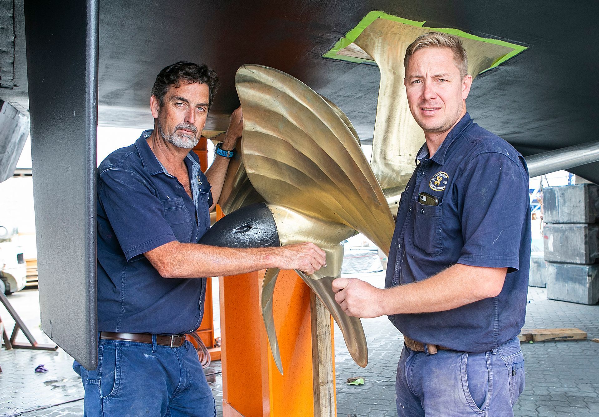Two men pose, holding a large boat propeller. They stand under a boat, in front of an orange surface.