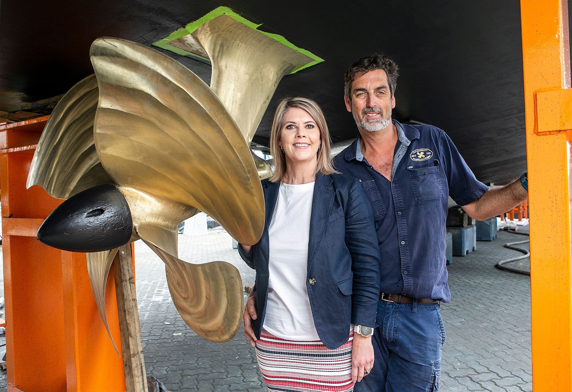 Woman and man stand beside a large ship propeller, under a black hull, in a shipyard.