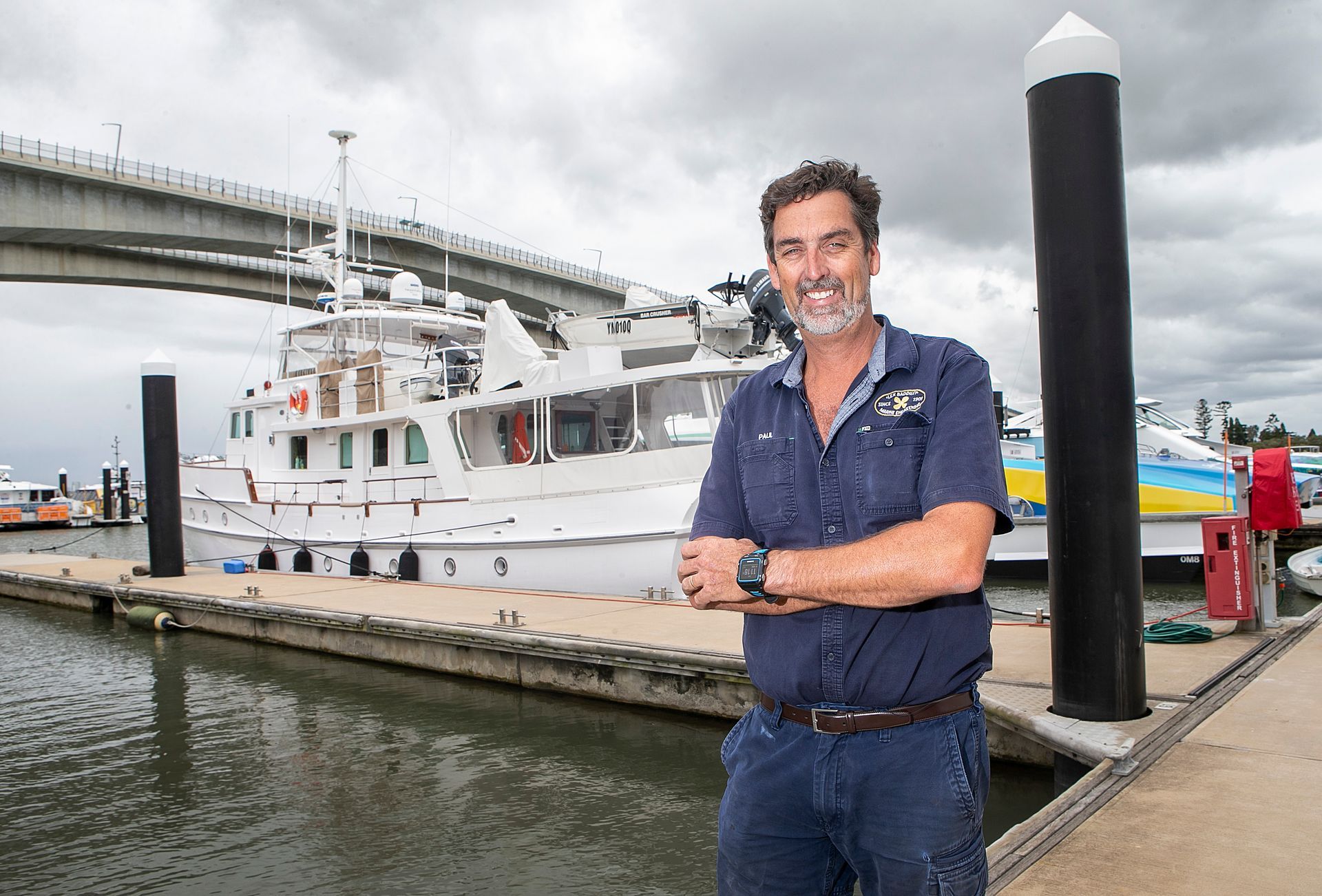 Man in blue shirt, arms crossed, standing on a dock with a large white boat and bridge in the background.
