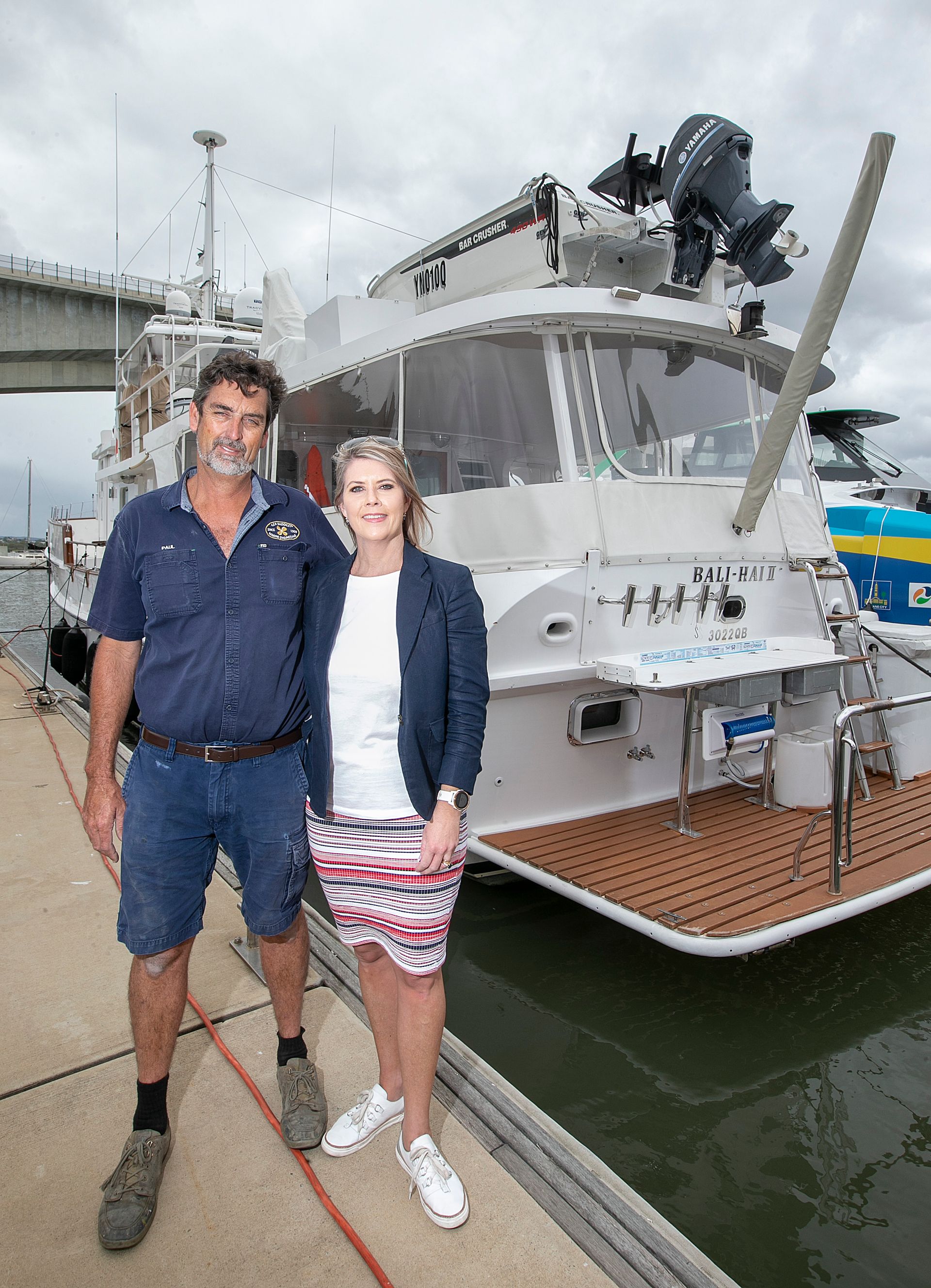 Man and woman stand on a dock next to a white boat. Man wears blue shirt, shorts. Woman wears jacket, shorts.