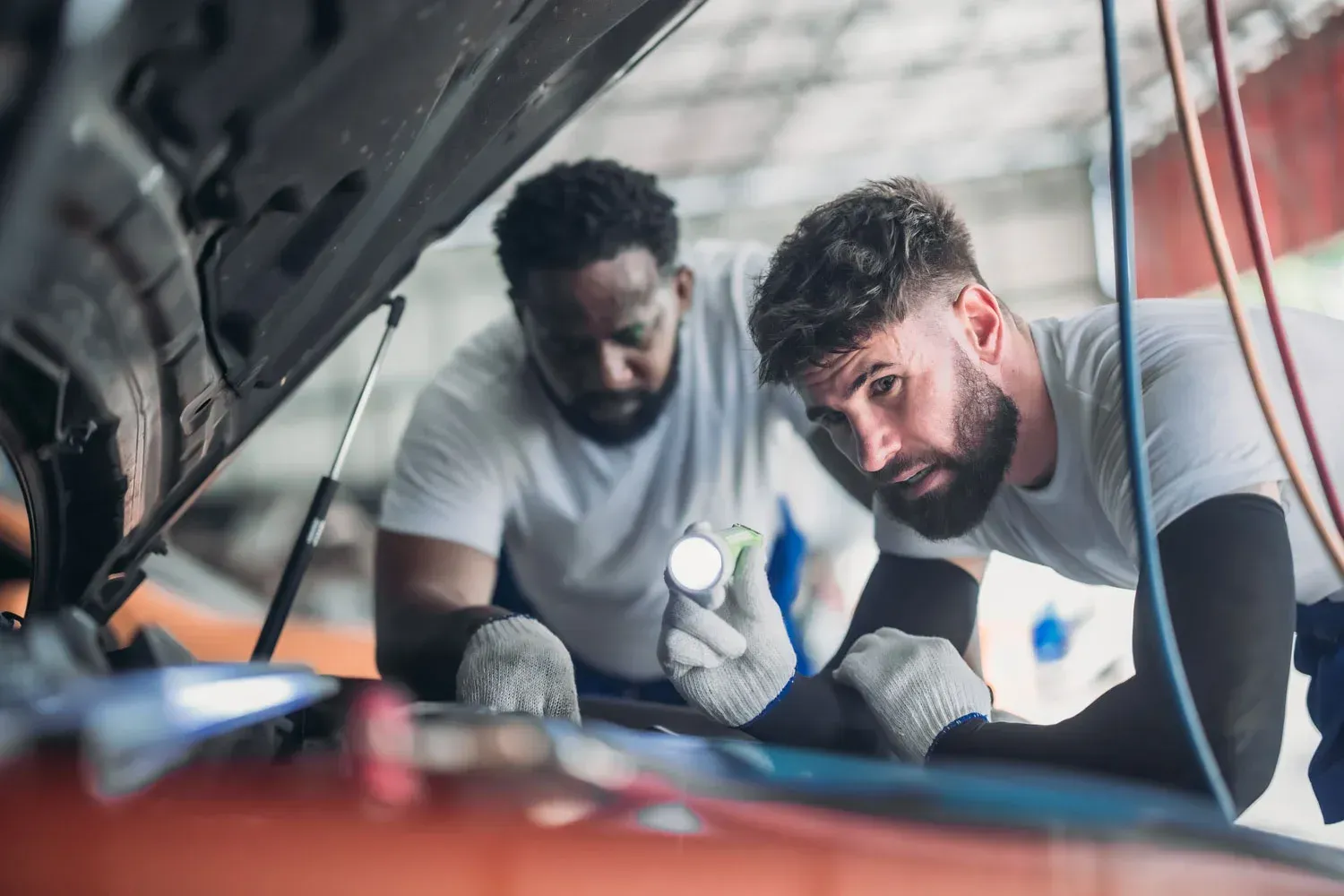 Two mechanics in a garage examine an open car engine, one using a flashlight to inspect the components.