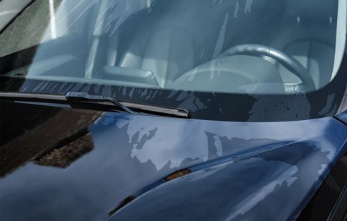 Close-up of a blue car hood showing significant clear coat peeling and bubbling, with the windshield and steering wheel.