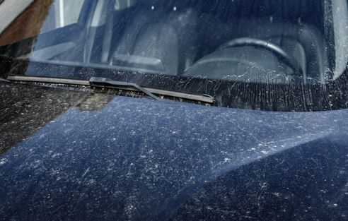 The hood and windshield of a dark, dusty car parked outdoors, showing heavy streaks and spots of grime on the surface.