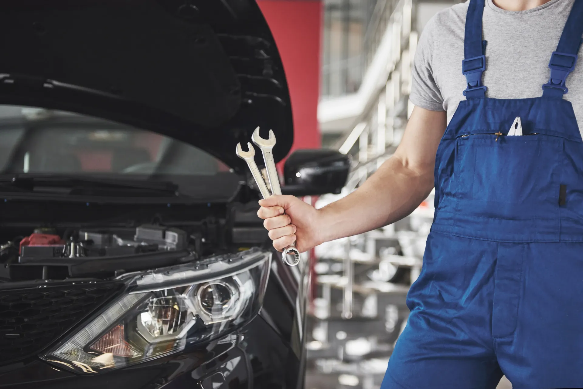 A mechanic in blue overalls holds two wrenches in front of a car with an open hood in a workshop.
