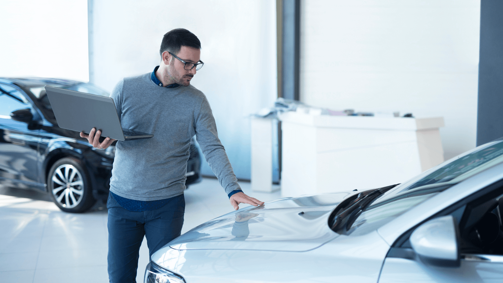 A person in a gray sweater holding a laptop stands in a car dealership, checking the hood of a silver car.