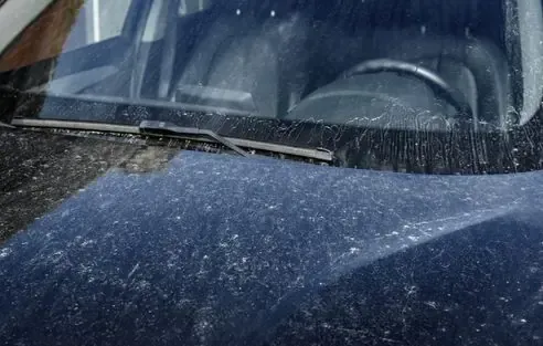 The hood and windshield of a dark, dusty car parked outdoors, showing heavy streaks and spots of grime on the surface.