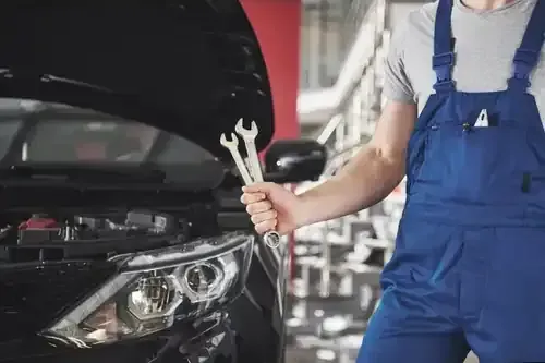 A mechanic in blue overalls holds two wrenches in front of a car with an open hood in a workshop.