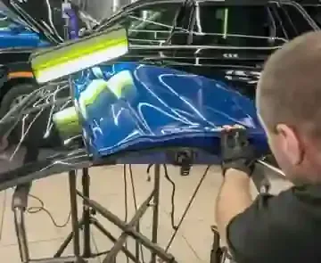 A technician uses a light board to perform paintless dent repair on a detached blue car hood in a workshop.