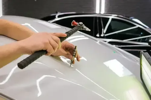 A technician uses a specialized tapping tool to perform paintless dent repair on a silver car roof in a workshop.