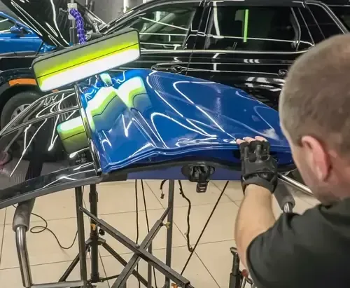 A technician uses a light board to perform paintless dent repair on a detached blue car hood in a workshop.