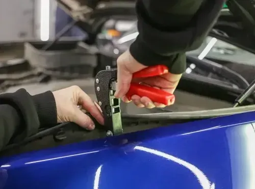 A person uses a red-handled professional dent puller tool to repair a small dent on a blue car hood.