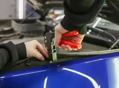 A person uses a red-handled professional dent puller tool to repair a small dent on a blue car hood.
