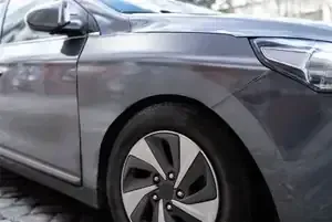 A close-up of a grey car's front passenger side, showing the wheel, fender, and headlight on a paved surface.