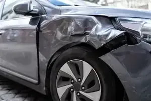 A damaged gray car with a crumpled front fender parked on a stone-paved surface.