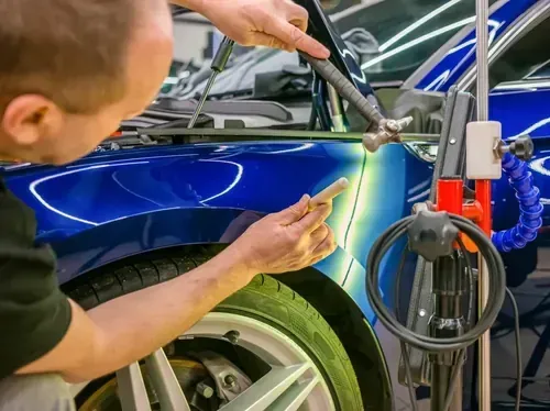 A mechanic uses a small hammer and a plastic punch to perform paintless dent repair on a blue car fender.