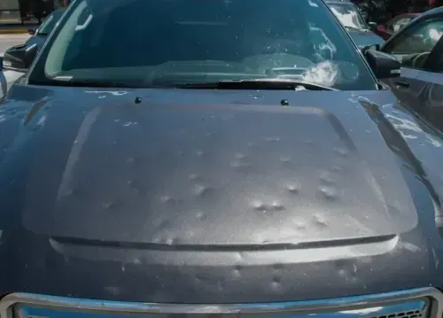 A dark gray car hood covered in numerous dents caused by hail damage.