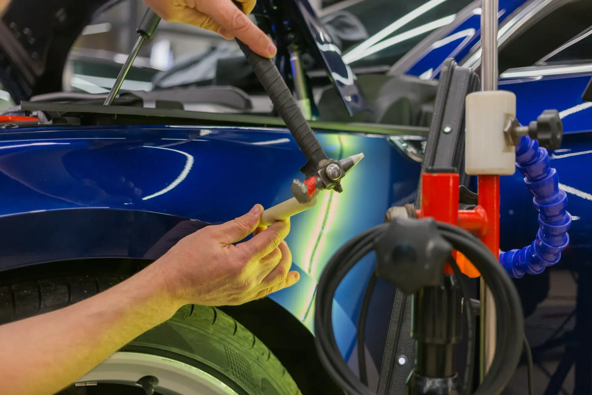 A mechanic uses a small hammer and a plastic punch to perform paintless dent repair on a blue car fender.