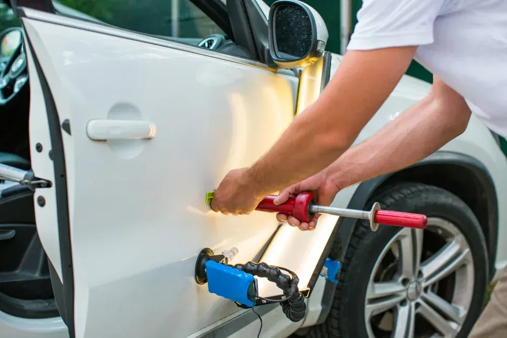 A mechanic uses a slide hammer tool to repair a dent on a white car door, illuminated by a specialized light board.