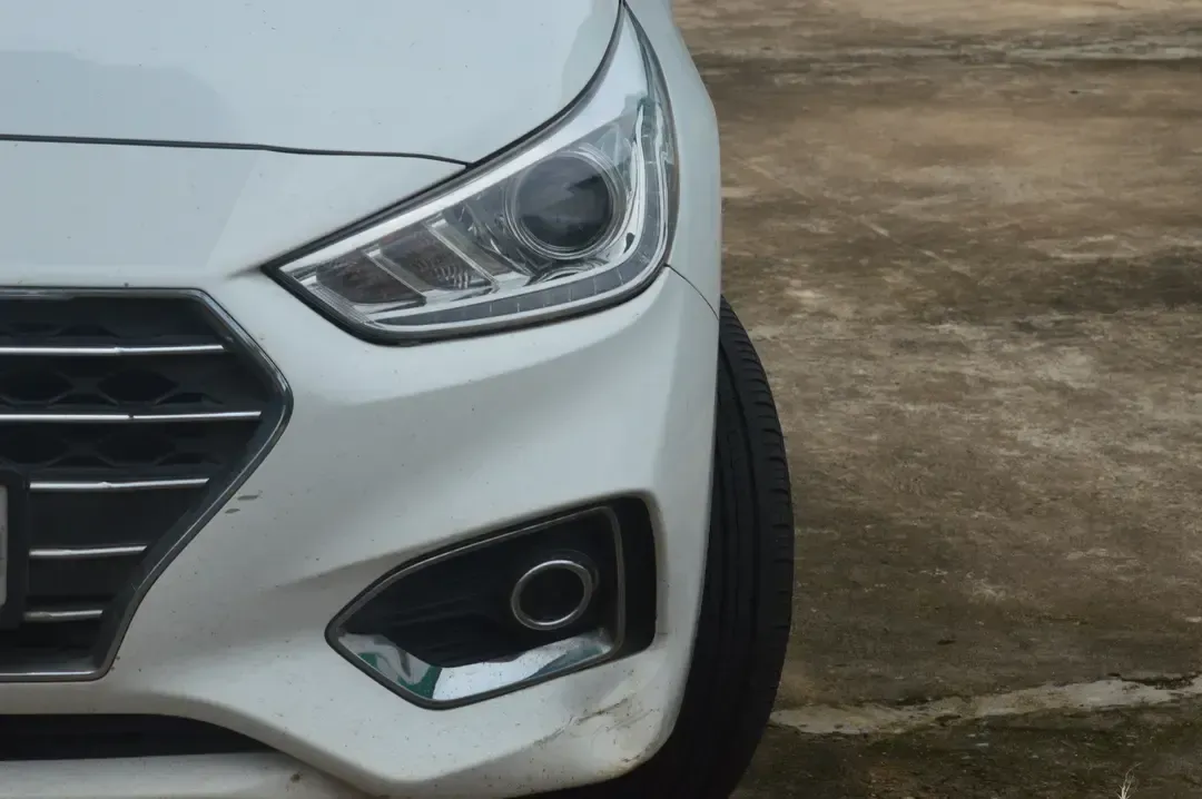 Close-up view of the front passenger-side corner of a white car, showing the headlight, fog light, and bumper damage.